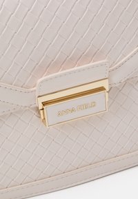 Handtasche in cremefarbenem Webmuster mit glatter Textur. Verfügt über einen goldfarbenen Verschluss und eine Markenplakette, die das strukturierte Design ergänzen.