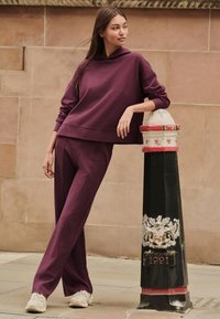 Hanorac scurt burgund și pantaloni asortați din țesătură moale. Modelul se sprijină de un stâlp stradal alb-negru cu accente decorative.