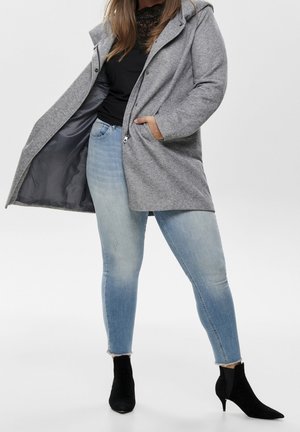 Persona indossa jeans skinny azzurri chiari, stivaletti neri con tacco, top nero e cappotto grigio con cappuccio aperto con la mano in tasca.