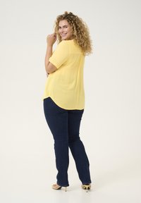 Blouse jaune à manches courtes avec une coupe décontractée, détail de pli au dos, associée à un jean bleu foncé légèrement évasé et des sandales à talons jaunes.