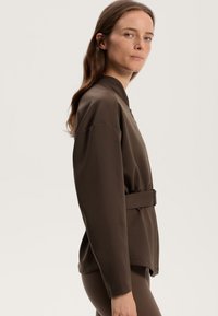 Brun jacka med hög krage, långa ärmar och bälte i midjan. Tillverkad av ett slätt tyg, med en minimalistisk design och en avslappnad passform.