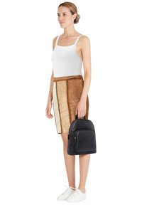 Black leather backpack with rounded shape, front zip pocket, smooth texture; worn with a fitted white tank top and a tan suede skirt.