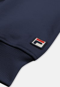 Marineblauwe stoffen mouw met geribbelde manchetten en een kleine rechthoekige patch met een abstract rood-zwart geometrisch ontwerp.