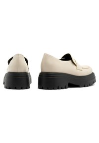 Beżowe skórzane buty na platformie z okrągłym noskiem, z dekoracyjną klapą i grubą czarną gumową podeszwą o teksturowanym wzorze.