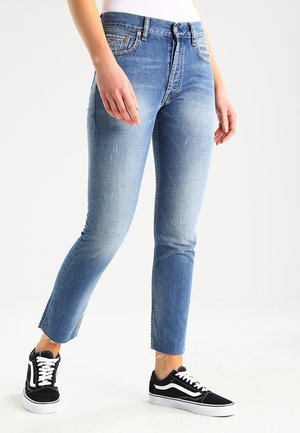 Person iført lyseblå falmede jeans med rå kanter og sort-hvide snørebåndssneakers, stående mod en ensfarvet lys baggrund.