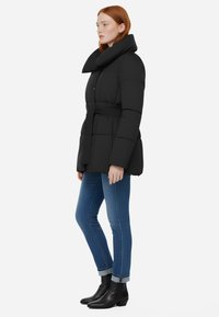 Giacca a vento nera con un ampio colletto ripiegato, vita con cintura e texture trapuntata, abbinata a jeans blu e stivaletti neri.