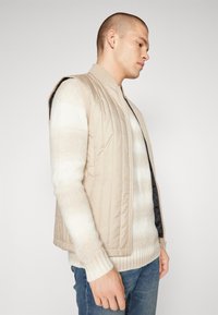 Gilet rembourré beige avec col montant, doté d'une fermeture éclair à l'avant, d'une texture matelassée et sans manches, porté par-dessus un pull en tricot de couleur claire.