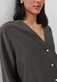 Chemisier gris foncé à col en V, à manches longues et au tissu lisse. Comporte un bouton blanc unique pour la fermeture.