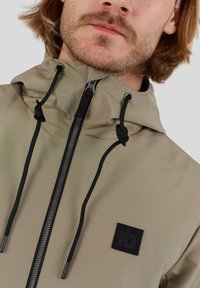 Wasserabweisende beige Jacke mit einem Reißverschluss, verstellbaren Kordeln und schwarzen Akzenten, die ein Logopatch auf der Brust trägt.