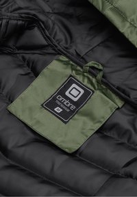 Étiquette en tissu vert avec l'inscription « ombre menswear M » cousue à l'intérieur de la doublure matelassée noire de la veste.