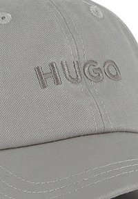 Gray baseball cap with stitched “HUGO” logo in matching thread, made of textured fabric. Features ventilation holes and curved brim.