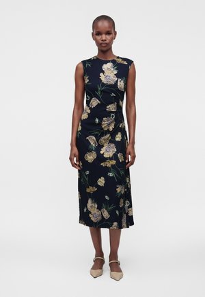 Femme debout, face à l'objectif, portant une robe midi bleu marine sans manches à motif floral jaune et des chaussures beiges à bout pointu, sur un fond uni.