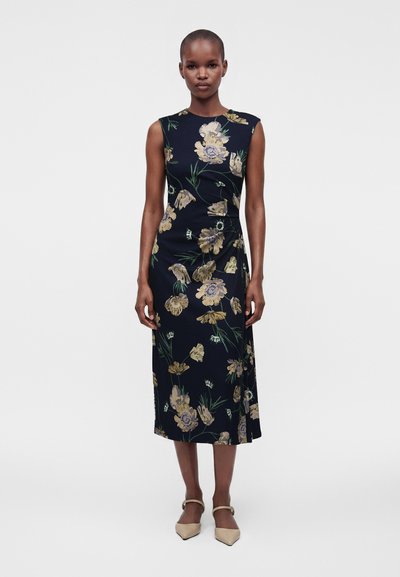 Γυναίκα στέκεται κοιτάζοντας μπροστά, φορώντας αμάνικο μπλε μίντι φόρεμα με κίτρινα λουλούδια και μπεζ μυτερές γόβες, σε απλό φόντο.
