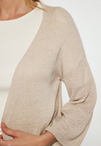 Beige stickad cardigan med lös passform och vida ärmar. Texturen är något genomskinlig med en fin väv, som är matchad med en krämfärgad top under.