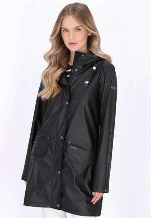 Kvinde med langt, bølget blondt hår iført en sort regnjakke med forlommer og knaplukning foran mod en ensfarvet hvid baggrund.