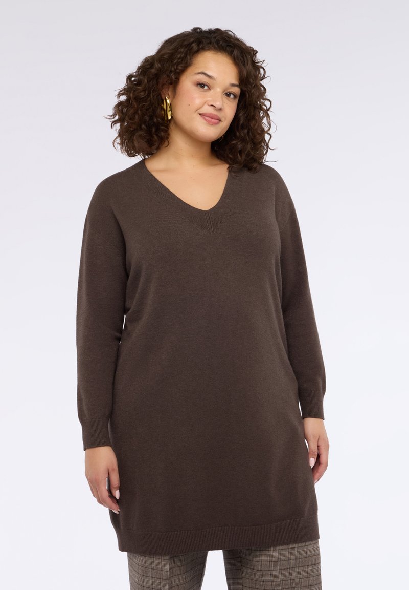 Vestito a maglia marrone con scollo a V e maniche lunghe, con polsini e orlo a coste. Indossato sopra pantaloni a quadri.