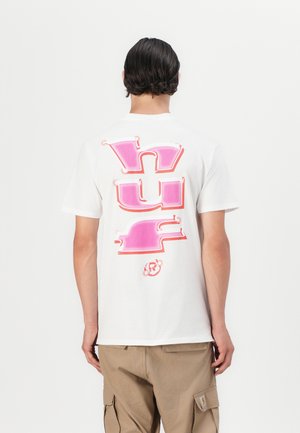 Bílý bavlněný tričko s velkým růžovým grafickým textem na zádech. Design obsahuje stylizované písmo a malé logo ve spodní části.