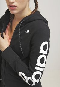 Haut de survêtement noir à zip avec un logo Adidas blanc contrastant sur la manche. Doté d'une capuche à cordon de serrage et d'une texture de tissu douce.
