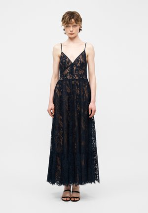 Giovane persona con un abito lungo in pizzo nero senza maniche, con spalline sottili e sandali neri aperti, in piedi davanti a uno sfondo bianco semplice.