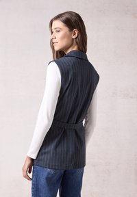 Marineblau gestreifter ärmelloser Weste mit Kragen und tailliertem Gürtel, getragen über einem weißen Langarmoberteil und kombiniert mit blauen Jeans.