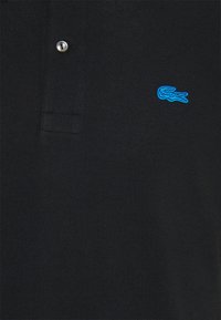Polo texturé noir avec deux boutons et un petit logo crocodile brodé bleu vif sur la poitrine.