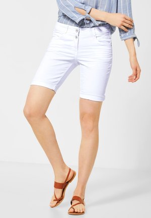 Frau trägt weiße knielange Shorts, blau gestreiftes langärmeliges Hemd und braune flache Sandalen, steht vor einem einfarbigen Hintergrund.