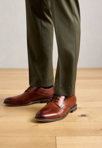 Chaussures habillées en cuir marron avec des lacets noirs, portées avec un pantalon vert foncé, se tenant sur un sol en bois clair.