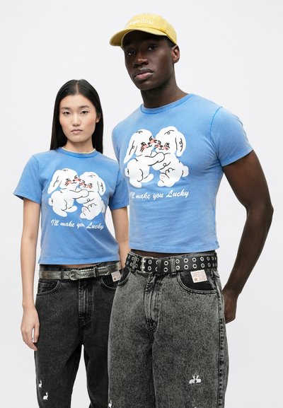 Blå t-shirts med kort ärm har en vit tecknad kanindesign med text. Kombinerade med lösa svarta jeans prydda med subtila detaljer och bälten.