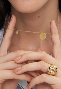 Złoty naszyjnik z dwoma zawieszkami: małym sercem i okrągłym dyskiem. Posiada delikatny łańcuszek o gładkiej fakturze. Ręce umiejscowione poniżej szyi.