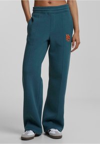 Pantalones de chándal teal con un corte relajado, cintura elástica, bolsillos laterales y logotipo bordado en naranja. Fabricados con una suave tela texturizada.