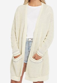 Luftig stickad cardigan i krämfärg med en lös passform, featuring två framfickor och ribbade mudd. Lager på en vit skjorta och denimshorts.