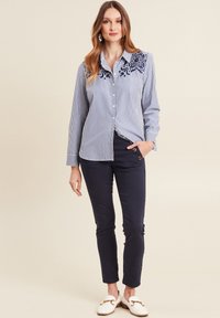 Chemise à boutons avec des rayures verticales bleu marine et blanches, ornée de motifs brodés bleu marine sur les épaules ; associée à un pantalon bleu marine et des mocassins blancs.