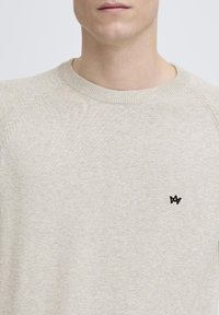 Beige genser med rund hals laget av teksturert strikket stoff. Har en liten svart logo på venstre bryst. Glatt, avslappet design med raglanermer.