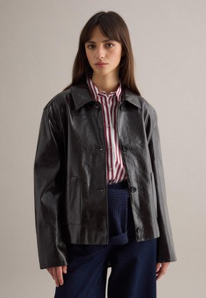 Jonge vrouw die een zwarte leren jas draagt over een rood-wit gestreept shirt en donkerblauwe broek, staand tegen een effen achtergrond.