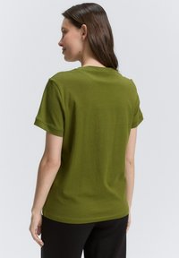 T-shirt en coton vert olive avec des manches courtes, col rond et coupe décontractée. Détails de couture à l'arrière, sans logos ni motifs visibles.