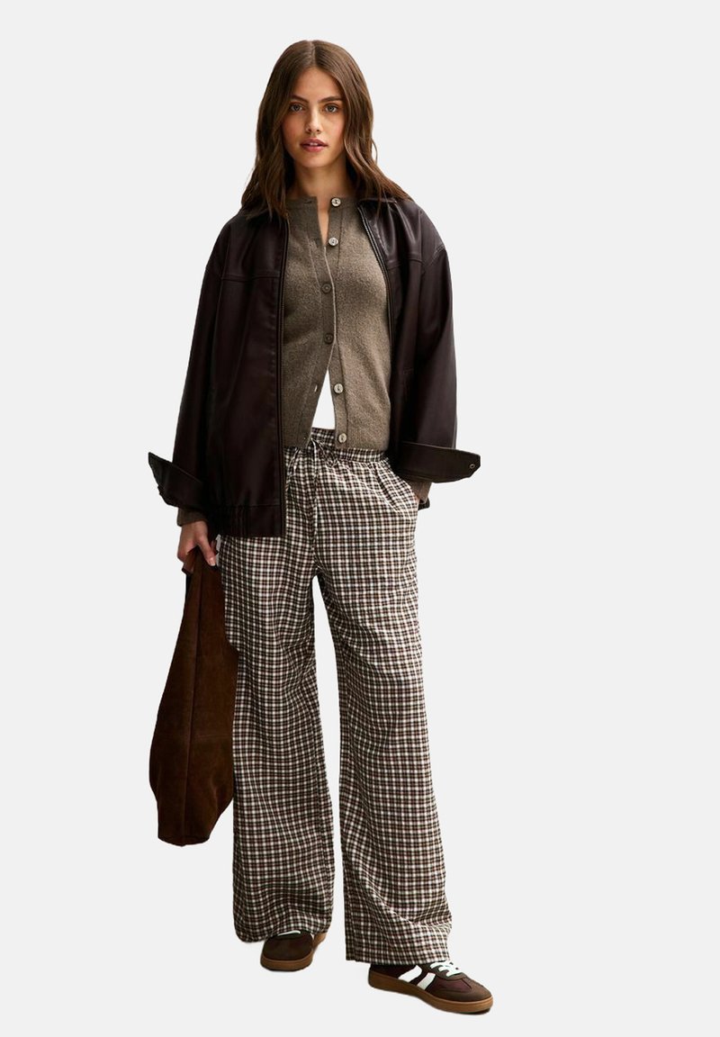 Chaqueta de cuero marrón sobre un cárdigan gris, combinada con pantalones de cuadros de gran tamaño. Zapatillas marrones con acentos blancos. Bolso tote marrón.