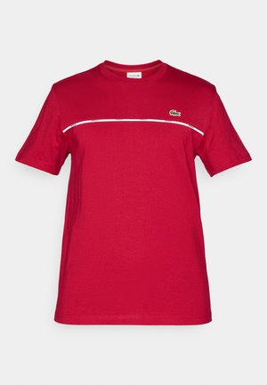 T-shirt en coton rouge à manches courtes, col rond, avec une rayure horizontale blanche portant du texte. Logo Lacoste en vert sur la poitrine gauche.
