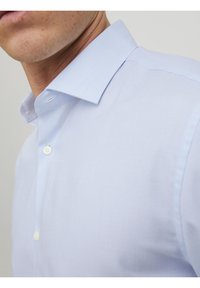 Detailní záběr muže v světle modré košili na knoflíky s pevně tvarovaným límcem, na jednobarevném pozadí.