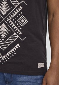 Chemise noire avec motif tribal géométrique blanc sur le côté, portée avec un jean bleu, montrant une partie du bras et de la main d'une personne.