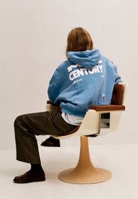 Felpa blu con testo "CENTURY", pantaloni grigi e scarpe marroni. Seduto su una sedia curva color crema con un accento in legno.