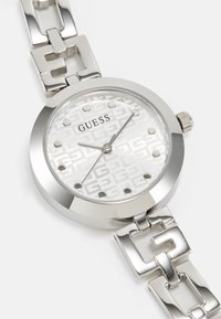 Zilveren ronde horloge met een gemarmerde witte wijzerplaat, metalen uurmarkeringen en een geschakelde armband. Merknnaam "GUESS" weergegeven op de wijzerplaat.