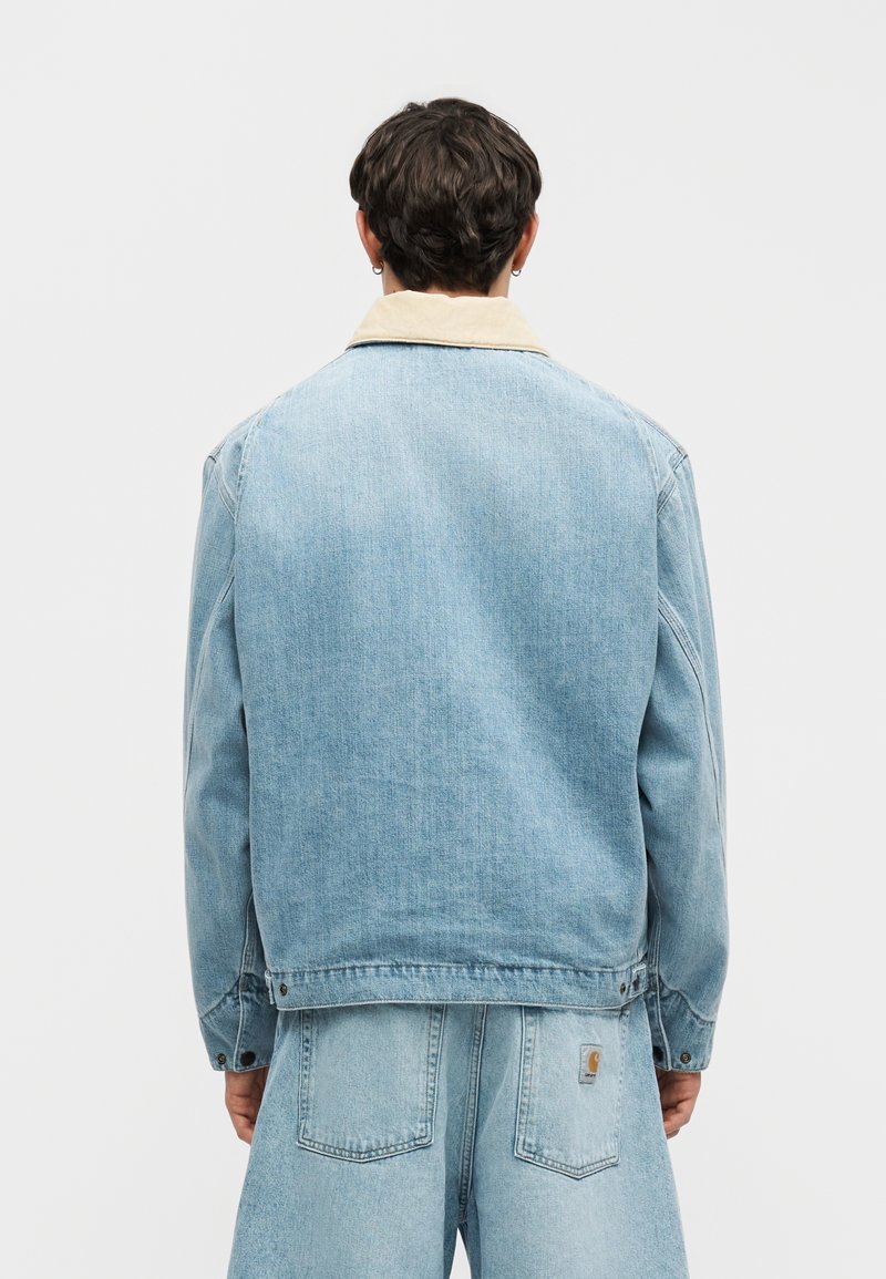 Persoon gezien van achteren, gekleed in een lichtblauw spijkerjasje met beige kraag en bijpassende spijkerbroek tegen een effen achtergrond.