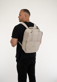 Beige ryggsäck med två axelremmar, framficka och logotyp, tillverkad av slätt tyg, visas bärande på en person bakifrån.