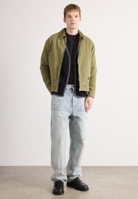 Young man wearing olive green zip-up jacket, black cardigan and t-shirt, loose light-wash jeans with distressing, and black shoes, standing indoors.