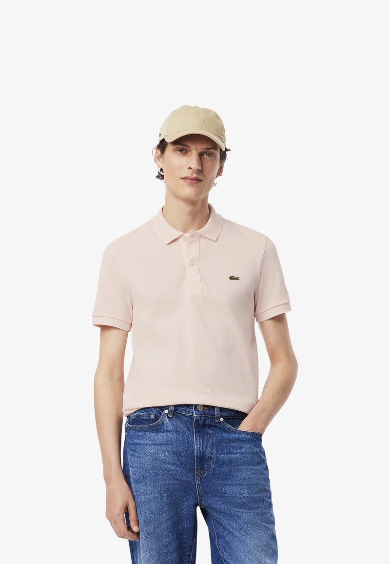 Ljusrosa pikétröja med korta ärmar och klassisk krage, kombinerad med blå denimjeans. Har en liten logotyp på bröstet.