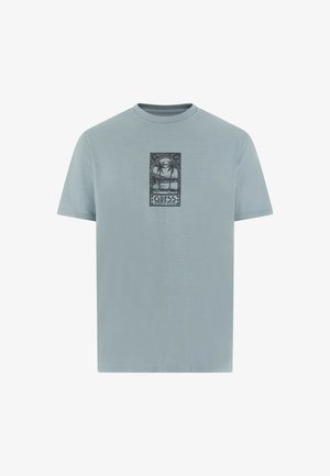 T-shirt en coton bleu clair à manches courtes, avec un graphique noir de palmiers et le texte "GUESS" au centre. Encolure ronde.