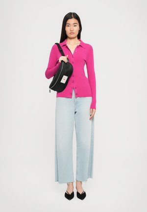 Frau in einem fuchsiafarbenen, geknöpften Cardigan, hellen, weit geschnittenen Jeans und schwarzen, spitz zulaufenden Schuhen, die eine schwarze Umhängetasche auf der Schulter trägt.
