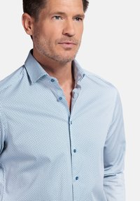 Chemise bleu clair à boutons avec un motif géométrique, dotée d'un col étalé et de boutons bleus, fabriquée dans un tissu lisse.