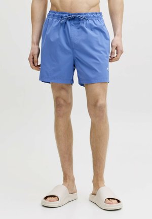 Man die blauwe zwemshort met trekkoord en beige slides draagt, staand tegen een effen witte achtergrond.