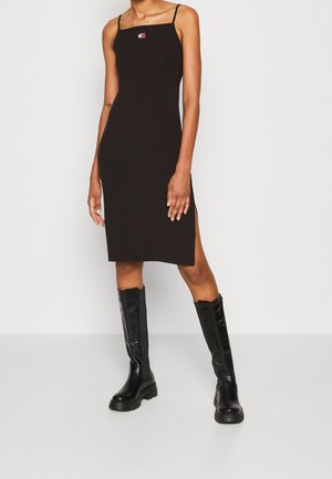 Frau trägt ein schwarzes ärmelloses knielanges Kleid mit Logo auf der Brust und einem seitlichen Schlitz, kombiniert mit schwarzen kniehohen Stiefeln.